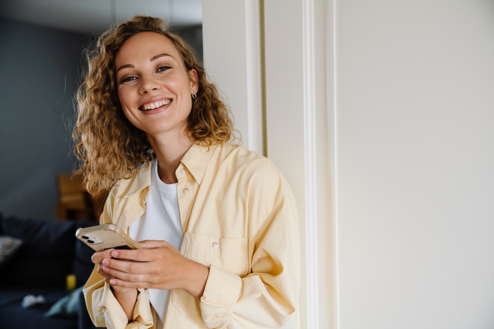 A woman with a bright smile holds a phone while standing indoors, showcasing her healthy teeth and radiant dental care - Dental Lab in Atlanta
