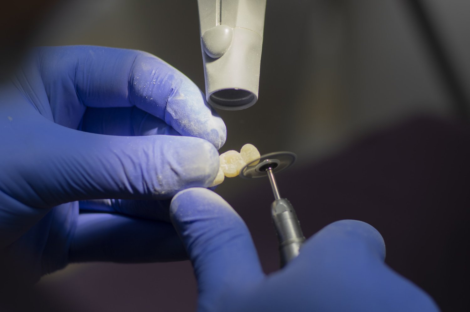 A dentist wearing blue gloves uses a dental tool to shape or polish a set of dental crowns or veneers under a magnifying scope - Dental Lab in Atlanta